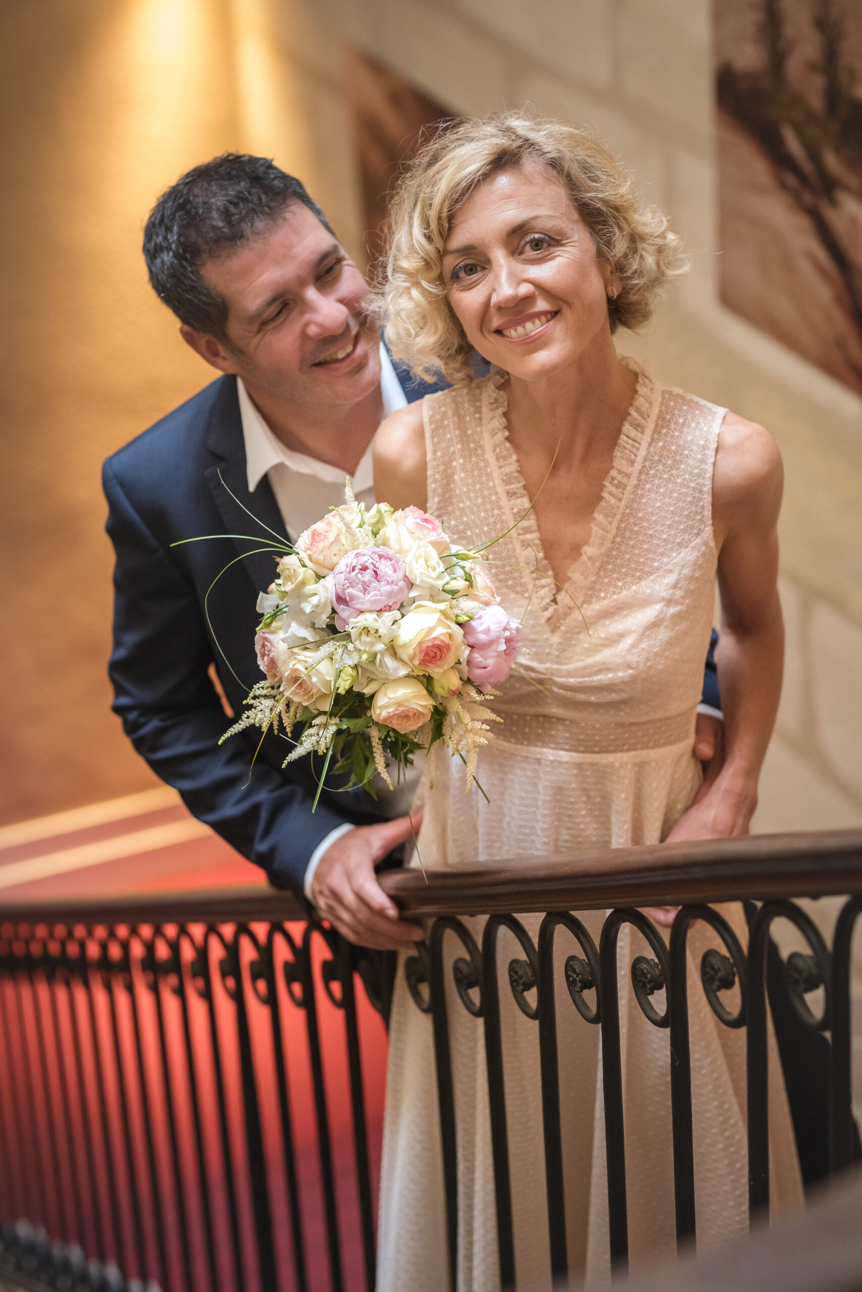 Mariés souriants appuyés sur une rambarde en fer forgé, bouquet de roses et pivoines