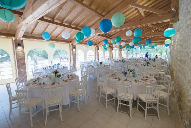 Salle de réception du Domaine des Charmilles, au Puy, sous une charpente décorée de lanternes turquoise et de tables rondes dressées