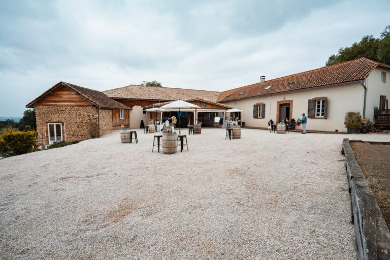 Cour en gravier du Belvédère du Domaine, à Tournay, avec ses tonneaux, ses parasols et ses bâtiments de ferme rénovés en galets et en bois