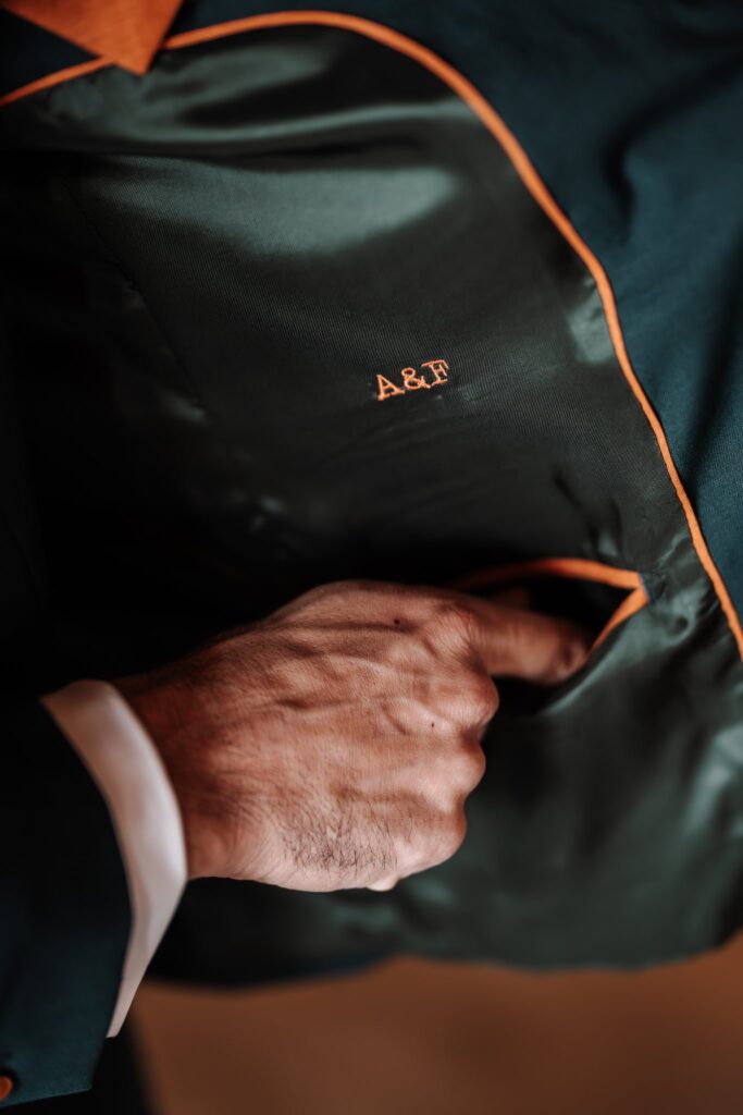 Une main glisse dans la poche intérieure d'une veste vert foncé, doublée d'un liseré orange et brodée des initiales A&F.