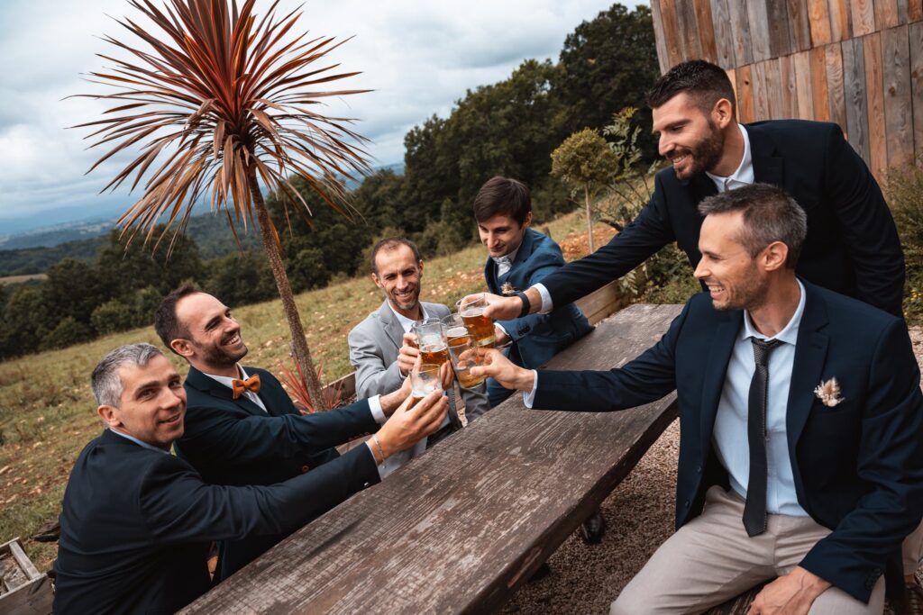 Cinq hommes en costume trinquent avec des verres de bière autour d'une table en bois, face aux collines et à un palmier.