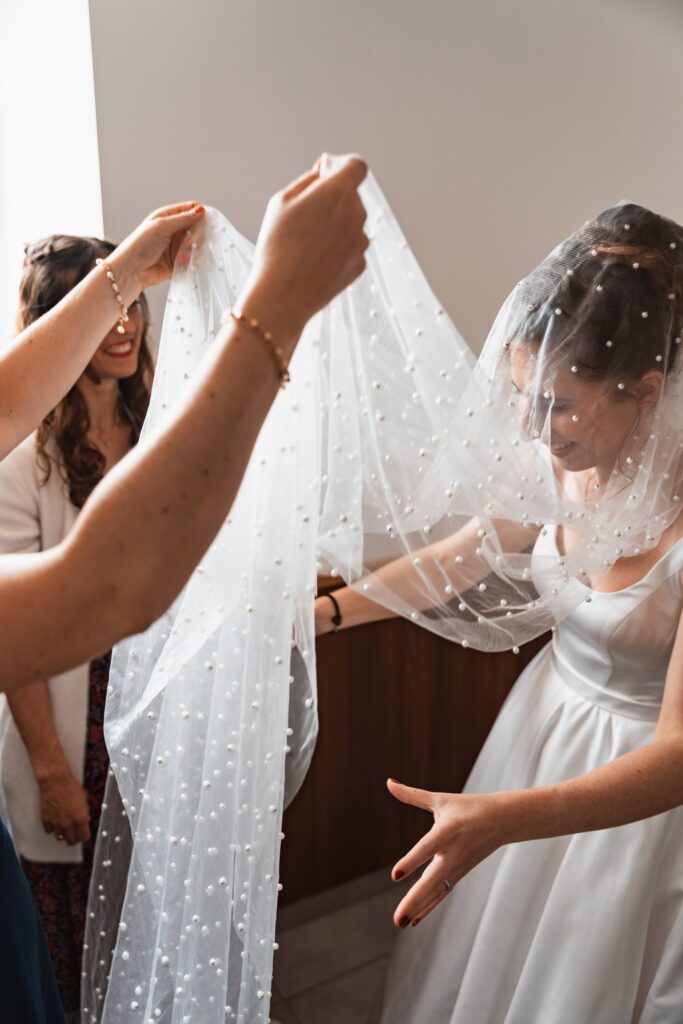 Deux mains écartent un long voile à perles pour le poser sur la tête de la mariée, en robe blanche à bretelles.