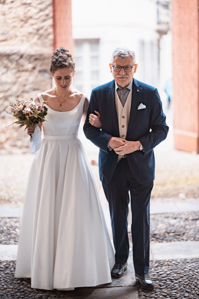 La mariée, son bouquet à la main, avance au bras d'un homme âgé en costume bleu à l'entrée de l'église.