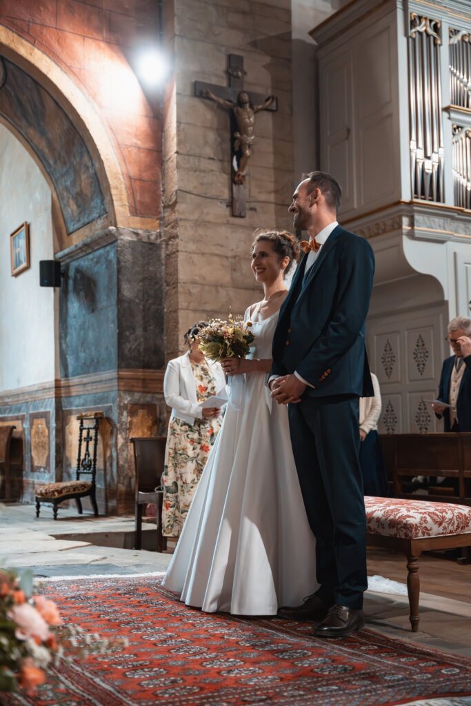 Les mariés se tiennent côte à côte devant l'autel de l'église, sur un tapis rouge, sous un grand crucifix.
