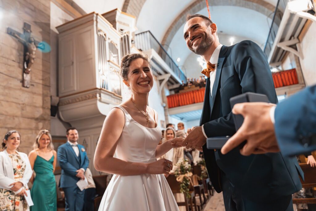 Dans l'église, la mariée sourit et le marié rit pendant l'échange des alliances, devant des invités debout.