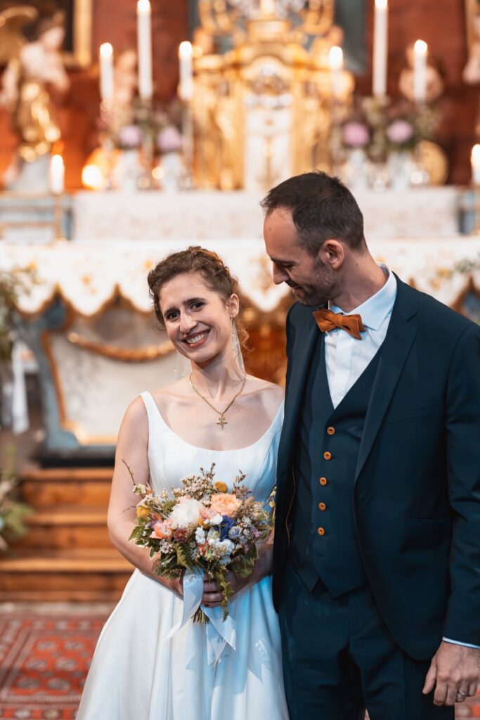 Devant l'autel doré de l'église, la mariée sourit à l'objectif avec son bouquet pendant que le marié baisse les yeux vers elle.