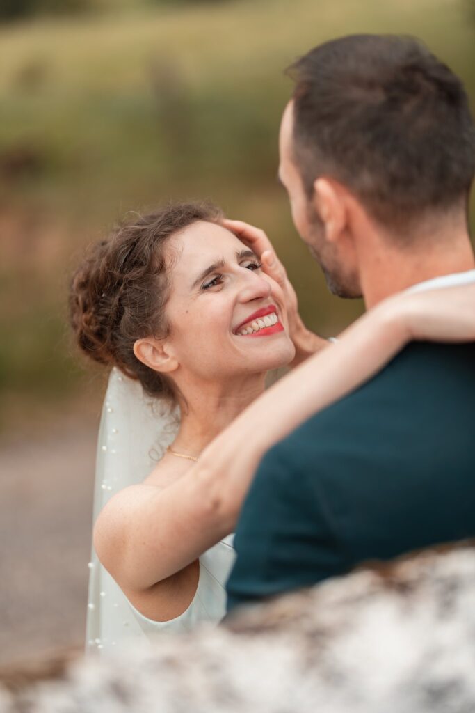 Gros plan sur le visage rieur de la mariée, la main sur la joue du marié qui la serre contre lui, sur fond de prairie floue.