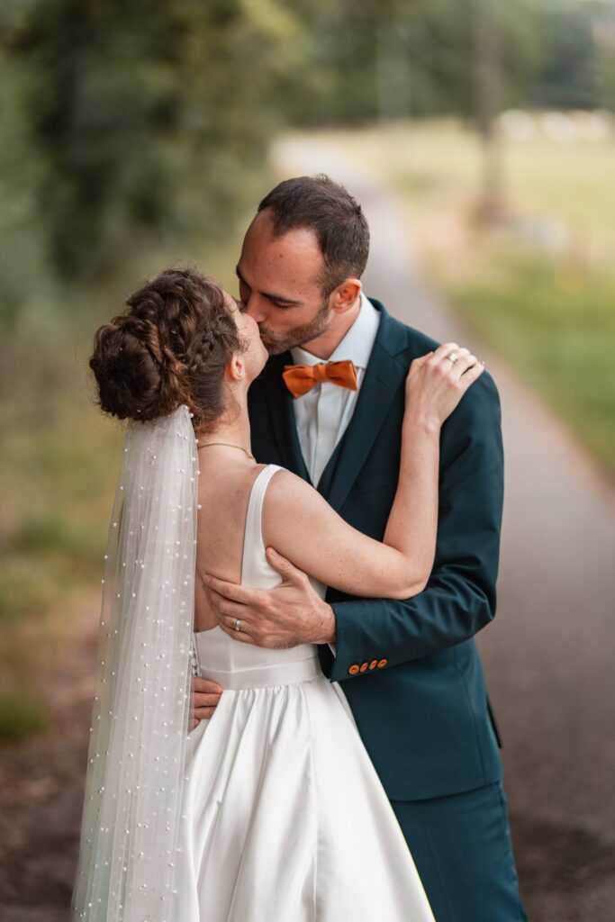 Le marié embrasse la mariée sur un chemin de campagne, son voile à perles retombant dans son dos.