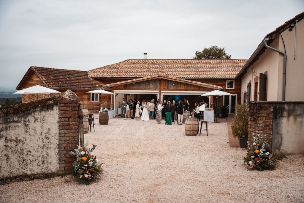 La cour du Belvédère du Domaine, un corps de ferme aux toits de tuiles, avec des bouquets de fleurs à l'entrée et des invités au fond.