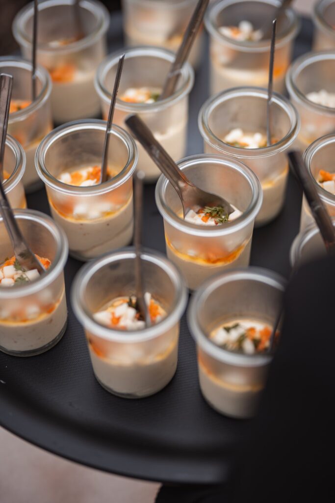 Un plateau de verrines garnies de crème, de légumes et d'une touche orange, tenues par de petites cuillères, pour le vin d'honneur.