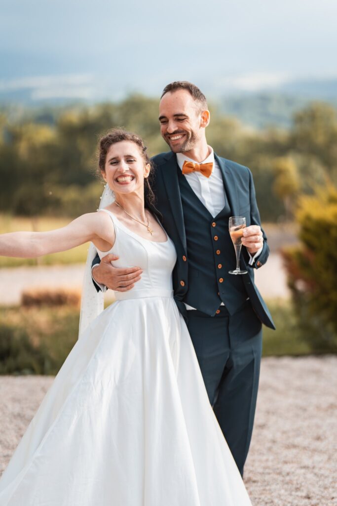 Dans la lumière dorée du soir, la mariée ouvre un bras en riant tandis que le marié, un verre à la main, l'embrasse sur la joue, avec des collines derrière eux.