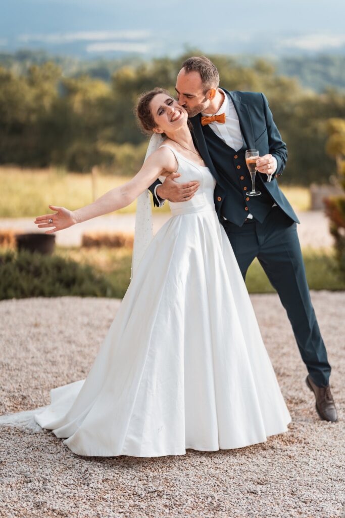 La mariée, bras ouvert, renverse la tête contre le marié qui l'embrasse, sur le gravier du Belvédère, avec les collines au loin.