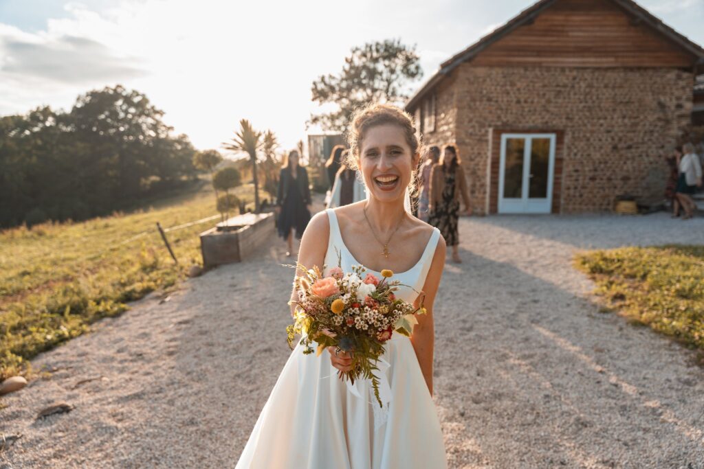 La mariée en robe blanche sourit en tenant son bouquet de fleurs sauvages, au soleil bas devant une bâtisse en pierre.