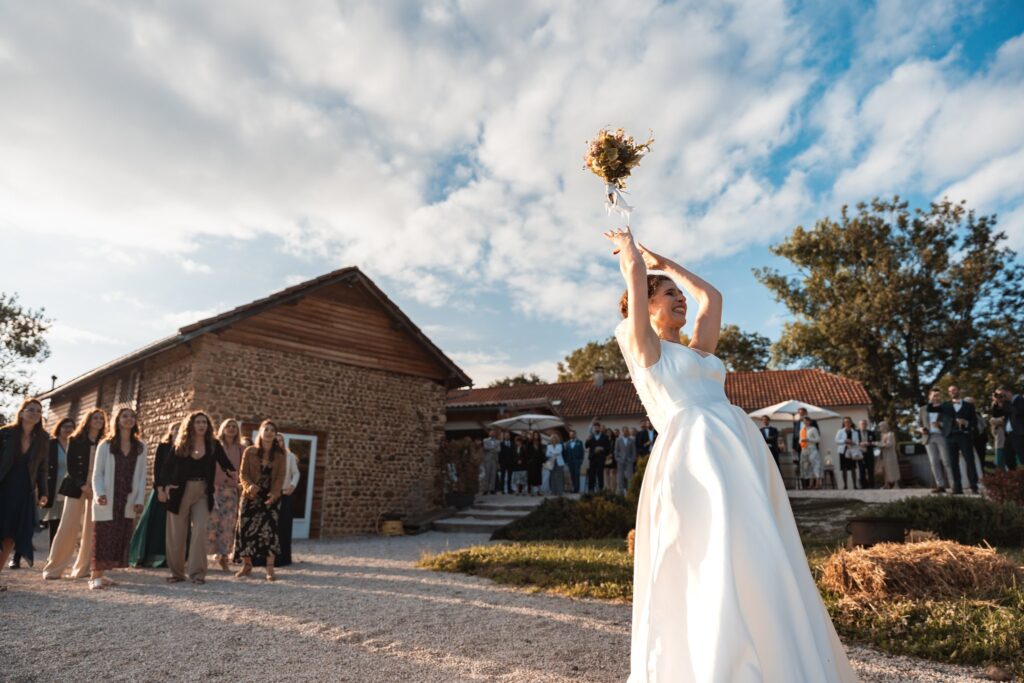 La mariée lance son bouquet vers le ciel bleu devant un groupe de personnes alignées près d'une grange en pierre.