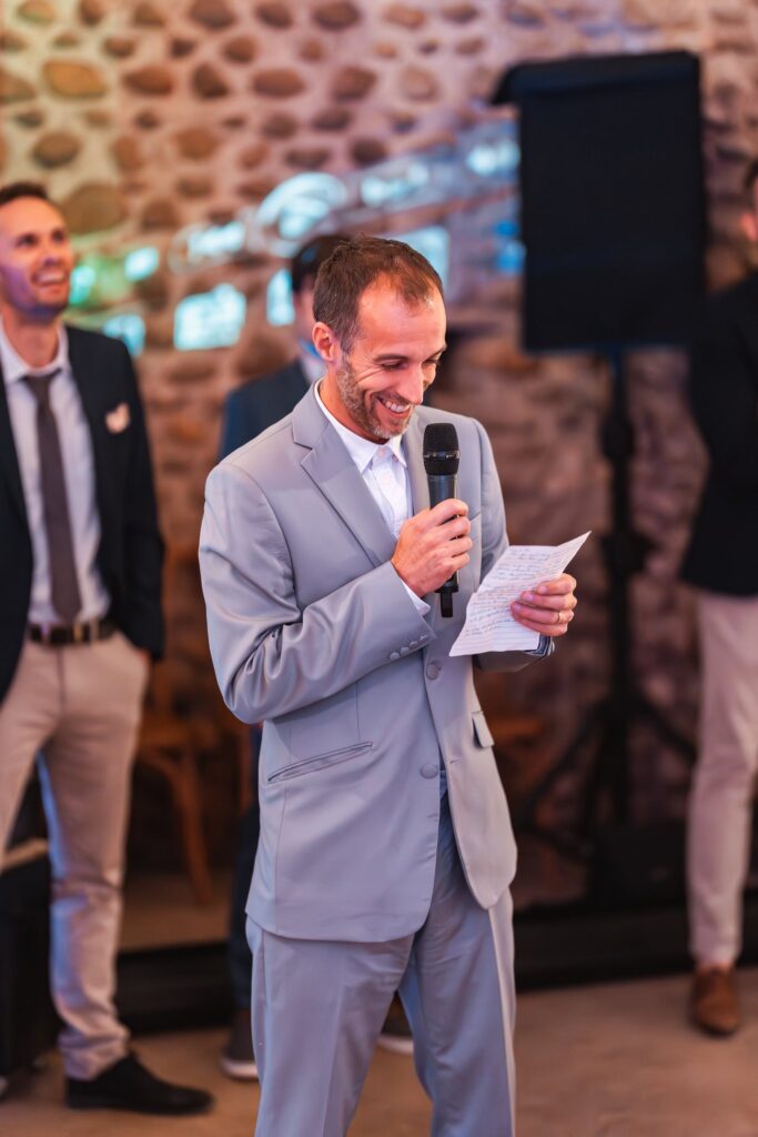Un homme en costume gris clair lit un discours au micro avec un grand sourire, devant un mur de galets.