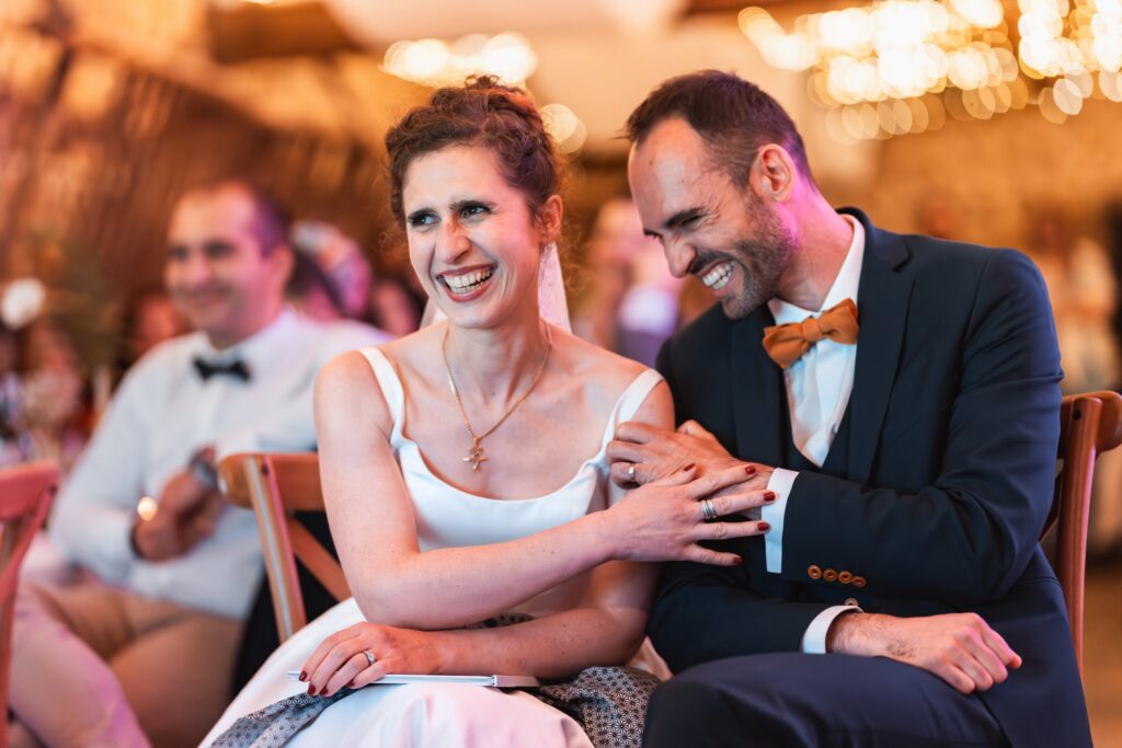 Les mariés sont assis côte à côte à table, penchés l'un vers l'autre et riant aux éclats, dans la lumière chaude de la grange.
