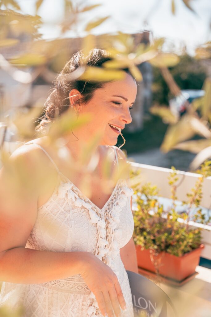 Une mariée en robe de dentelle sourit sur un balcon fleuri, à travers le feuillage.