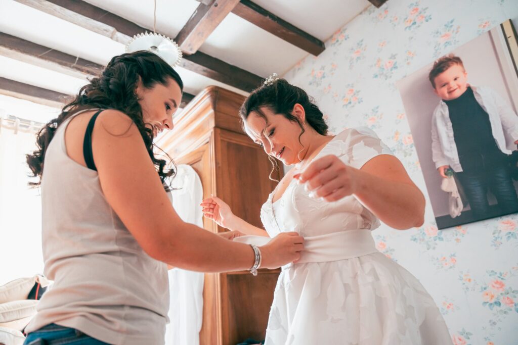 Une femme ajuste la ceinture de la robe de la mariée dans une chambre aux murs tapissés de fleurs.