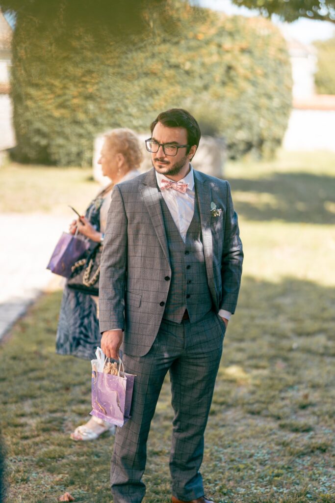 Le marié en costume gris et lunettes attend debout sur une pelouse ensoleillée.