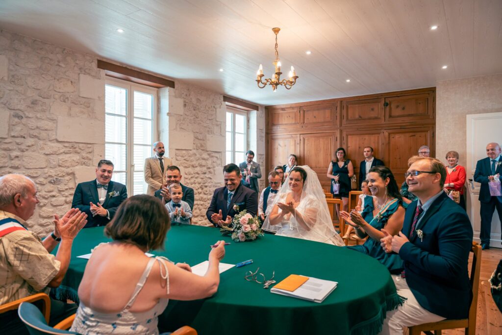 Dans une salle aux murs de pierre, des invités applaudissent autour d'une table de signature à la nappe verte.