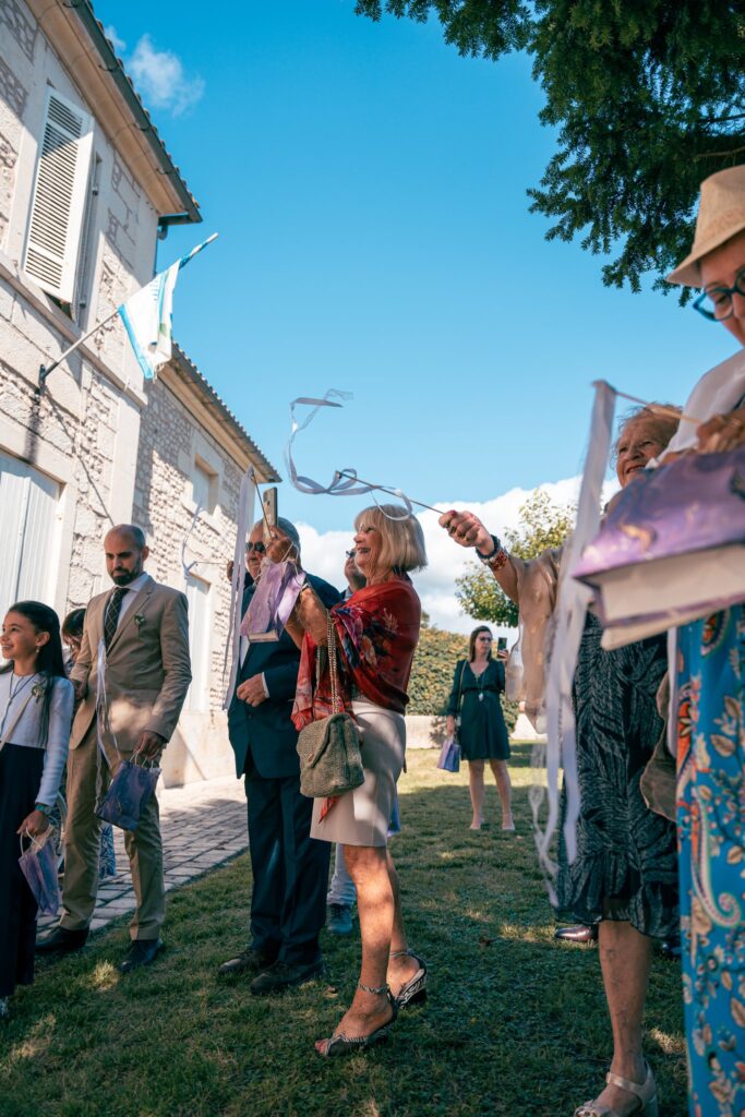 Des invités lèvent des rubans sous un ciel bleu devant un bâtiment en pierre.