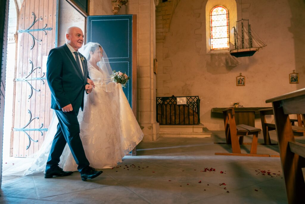 Un homme âgé accompagne la mariée voilée à l'entrée d'une église de pierre.
