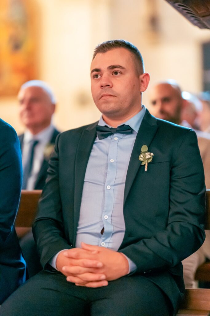 Le marié en costume sombre et nœud papillon, assis dans l'église, les mains jointes.