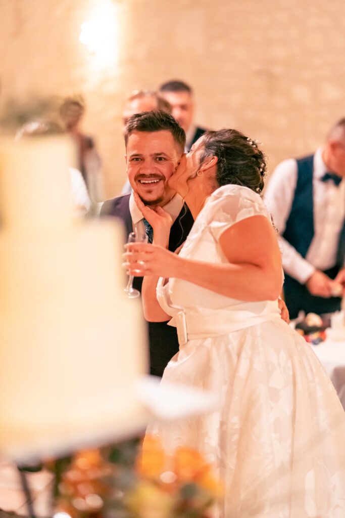 La mariée embrasse le marié sur la joue près du gâteau, dans une salle éclairée chaudement.