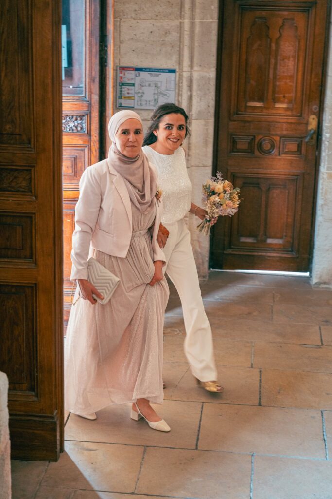 Deux femmes en tenue claire, dont l'une tient un bouquet, passent une grande porte en bois.