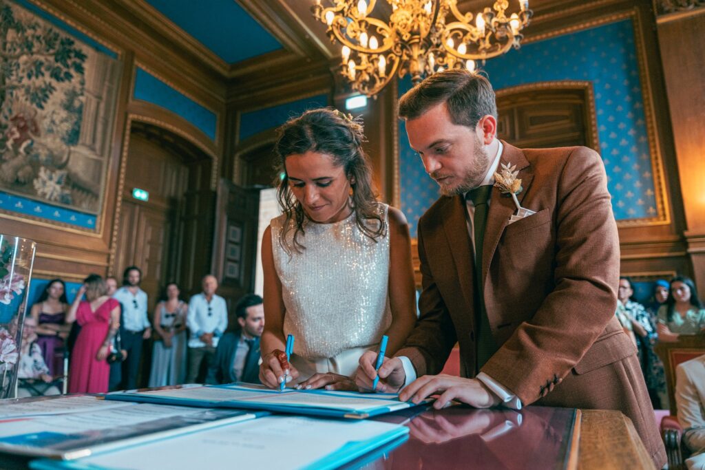 La mariée et le marié signent le registre, penchés côte à côte sur la table de la salle des mariages.