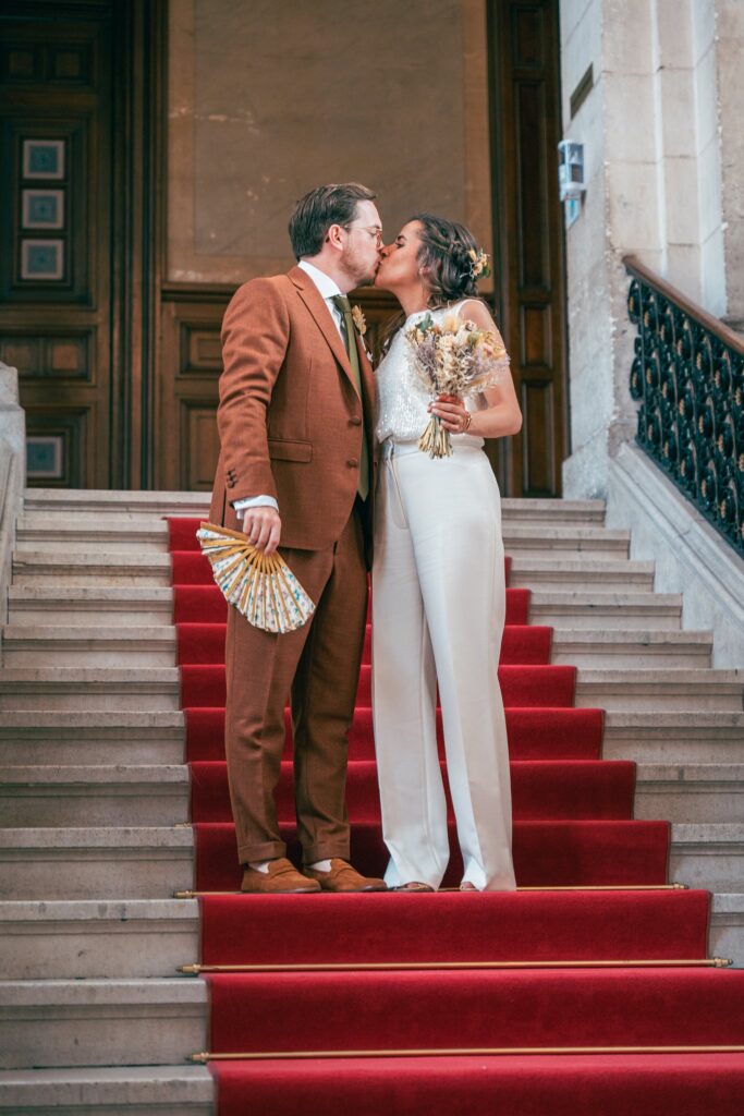 Le marié en costume rouille et la mariée en combinaison blanche s'embrassent sur un escalier recouvert d'un tapis rouge.