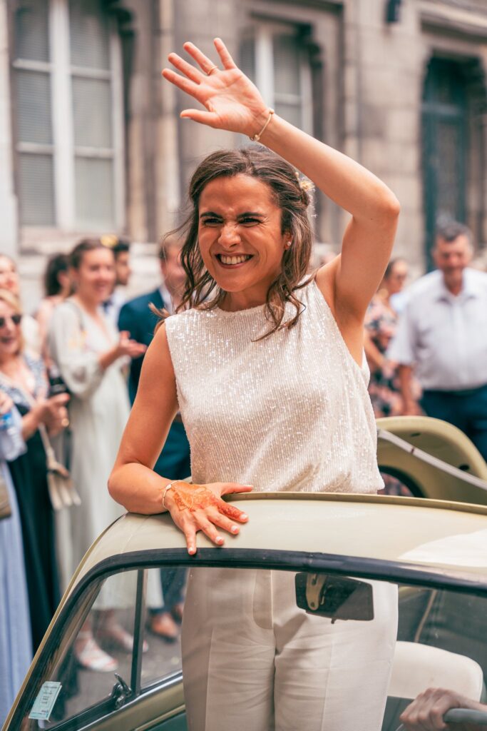 La mariée salue la foule d'un grand geste de la main, debout contre la portière d'une voiture ancienne, le sourire plein.