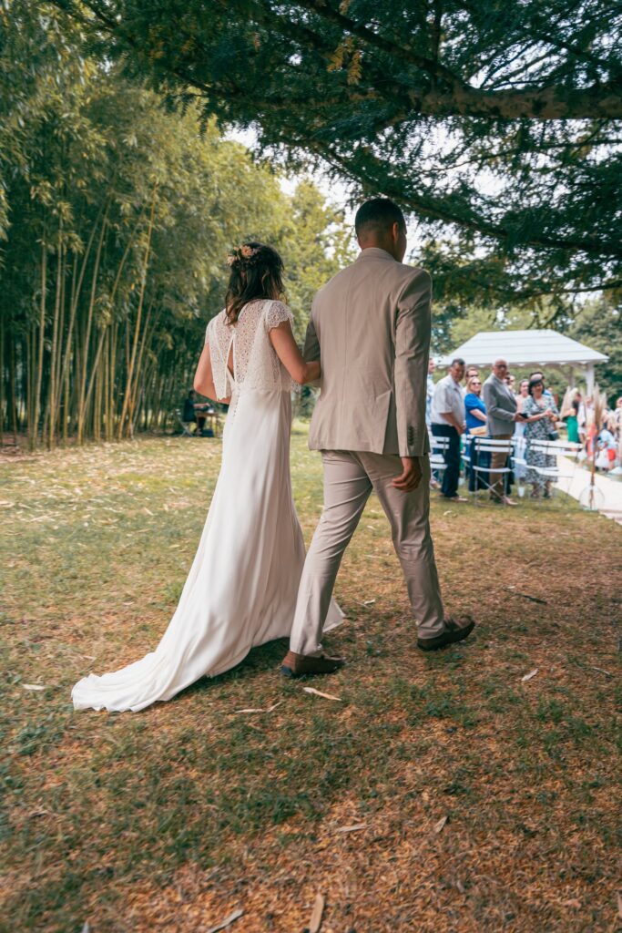 De dos, la mariée en robe longue avance sur l'herbe au bras d'un homme en costume clair, entre les arbres.