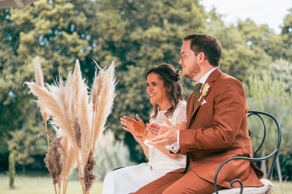 Assis côte à côte sur une chaise en fer forgé, le marié et la mariée applaudissent, avec des herbes de la pampa à leurs côtés.