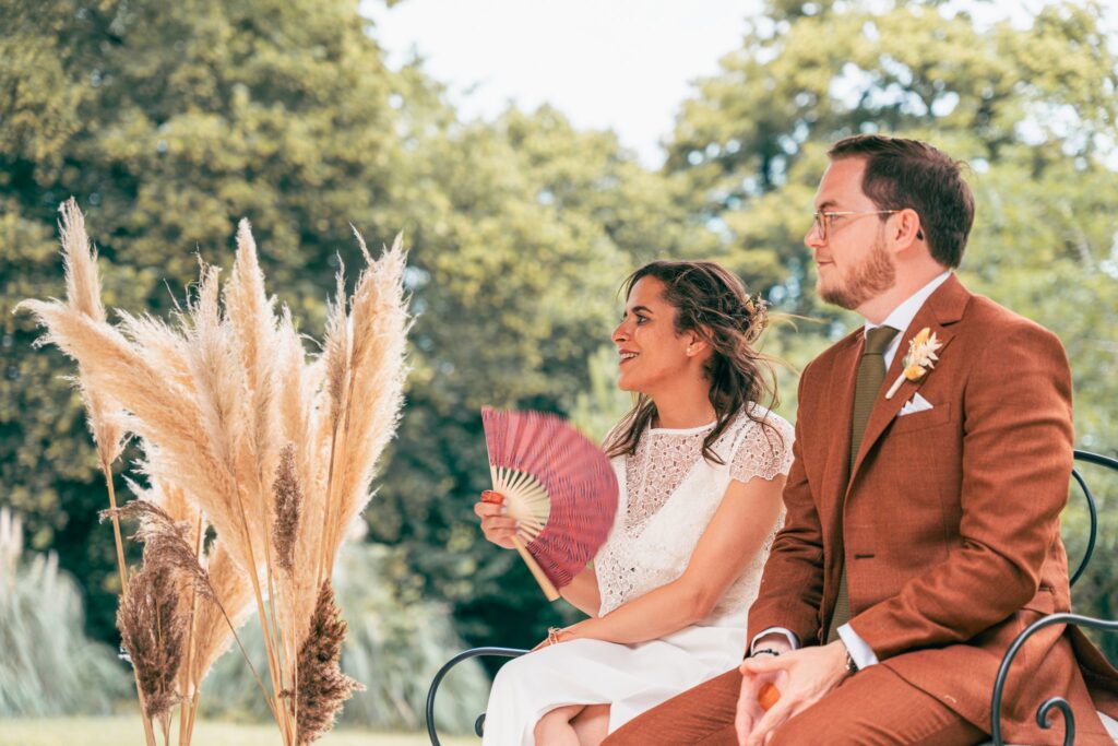 Le couple assis sous le kiosque blanc sourit, la mariée tenant un éventail rose, entre des bouquets de pampa.