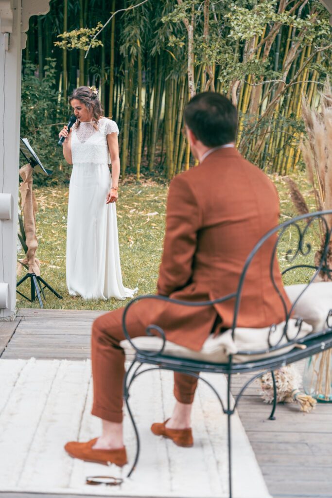 La mariée, debout dans l'herbe, parle au micro, face au marié assis de dos sur une chaise en fer forgé.