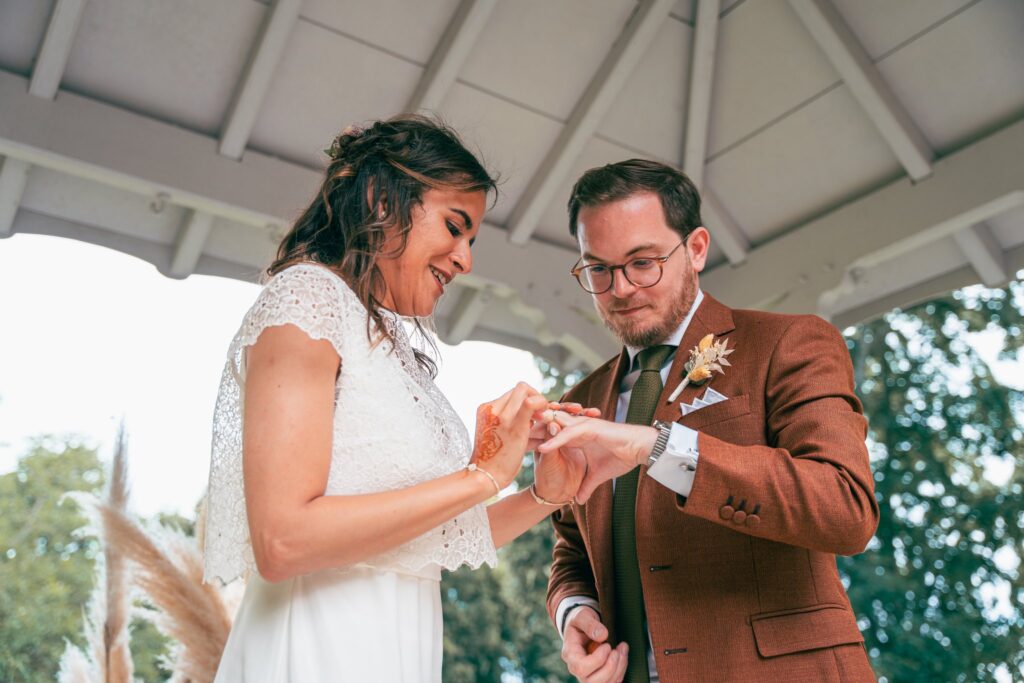 Sous le kiosque, la mariée passe l'alliance au doigt du marié, les mains au premier plan.