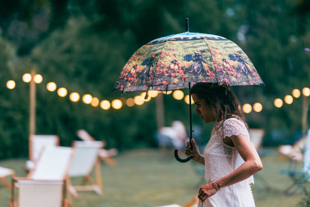 La mariée en robe blanche marche sous un parapluie à motifs, sous des guirlandes lumineuses, au-dessus de transats.