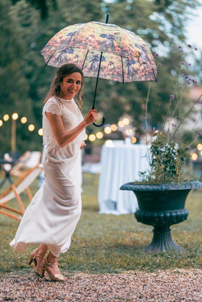 La mariée en robe longue blanche avance sur le gravier, un parapluie fleuri à la main, près d'une grande vasque.
