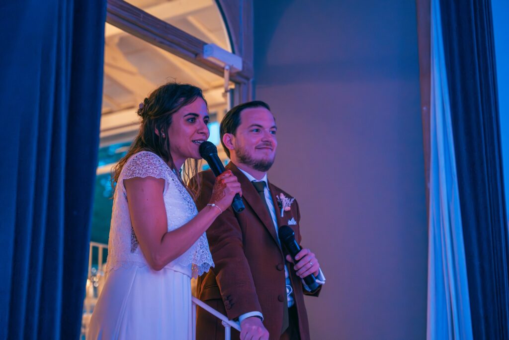 La mariée et le marié, chacun un micro à la main, prennent la parole dans la lumière bleutée de la salle de réception.