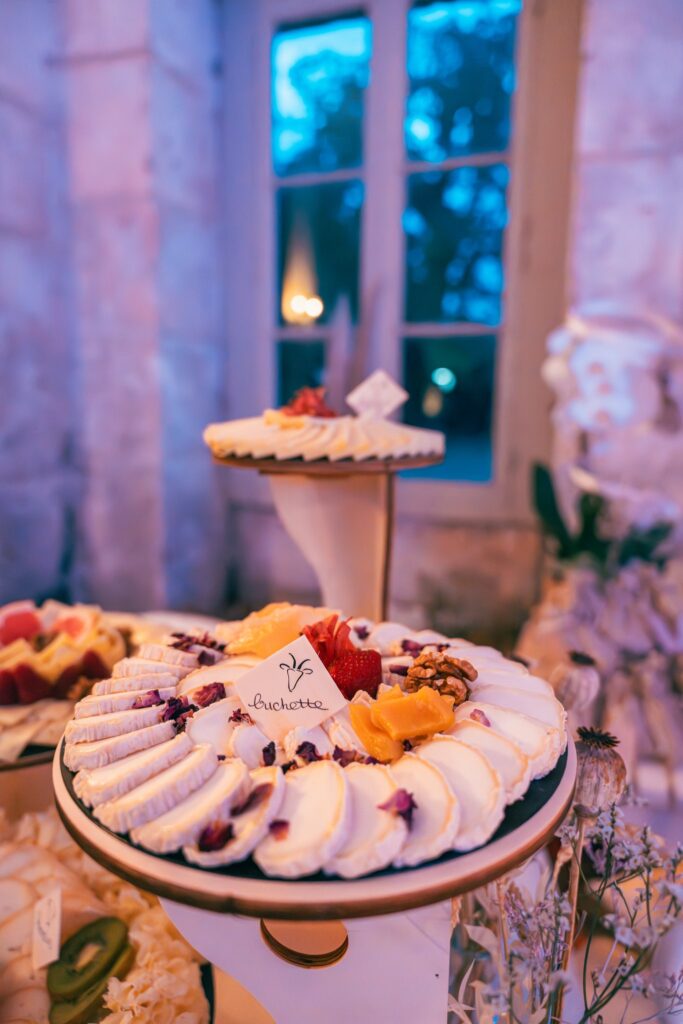 Un plateau de fromages décorés de fruits sur une table de buffet, devant une fenêtre aux reflets bleutés.