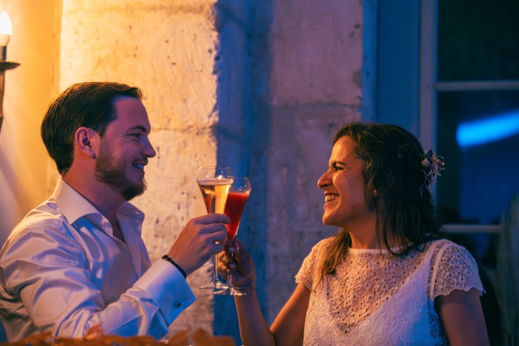 La mariée et le marié trinquent en riant, un verre à la main, dans la lumière chaude de la soirée.