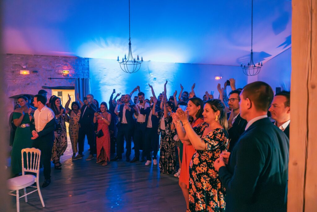 Dans une salle éclairée de bleu, des invités lèvent les bras et applaudissent, certains photographient la scène.