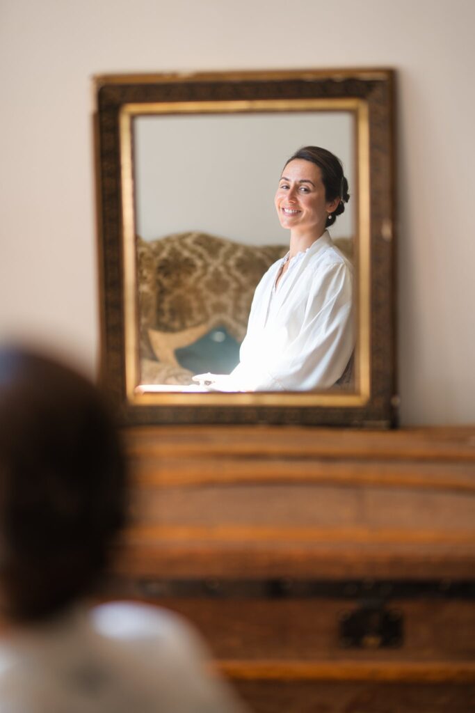 Dans un miroir ancien, une femme en peignoir blanc attend les préparatifs, le visage serein.