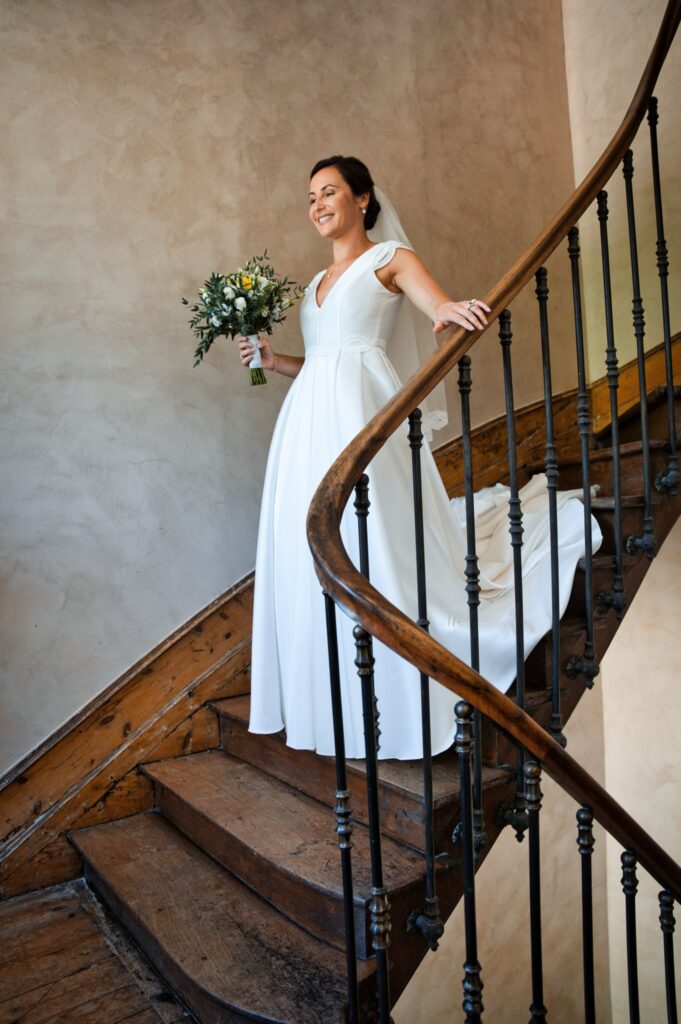 Une femme en robe blanche et voile tient un bouquet, debout dans un escalier en bois à rampe de fer forgé.