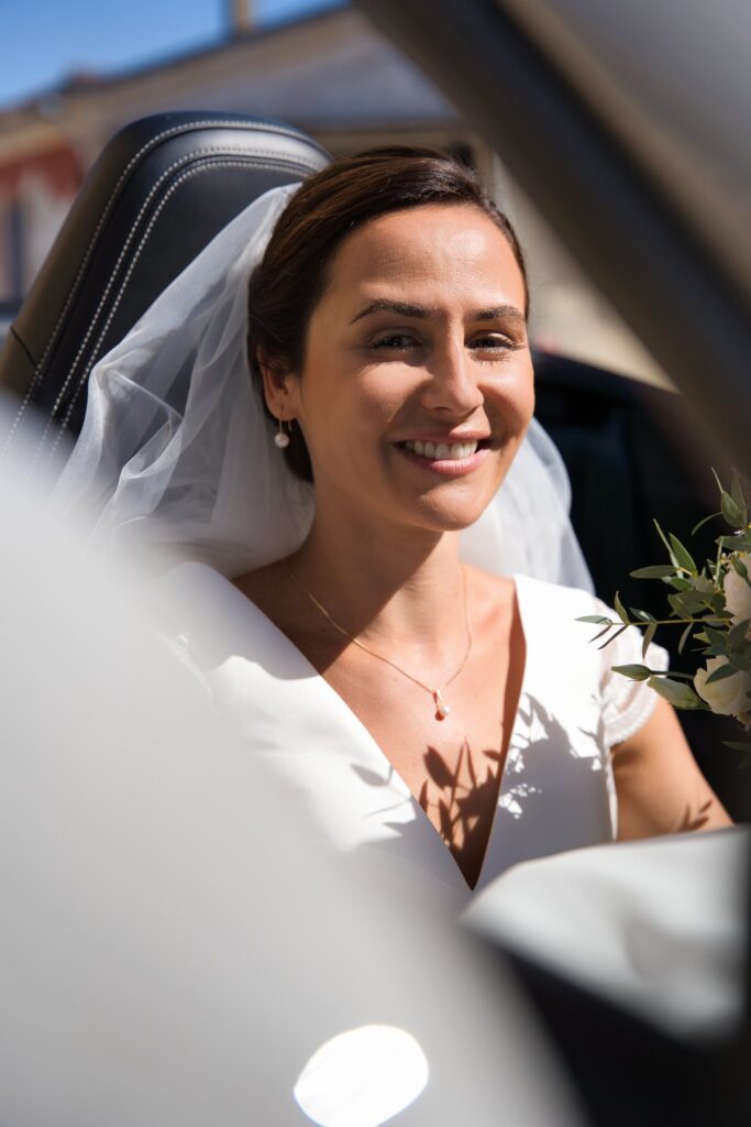 Une mariée en voile sourit à l'arrière d'une voiture, un bouquet de feuillage à la main.
