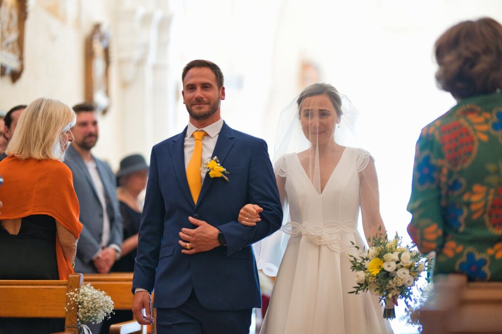 Dans l'allée centrale de l'église Saint-Vivien, la mariée avance sous son voile, au bras d'un homme en costume bleu à cravate jaune.