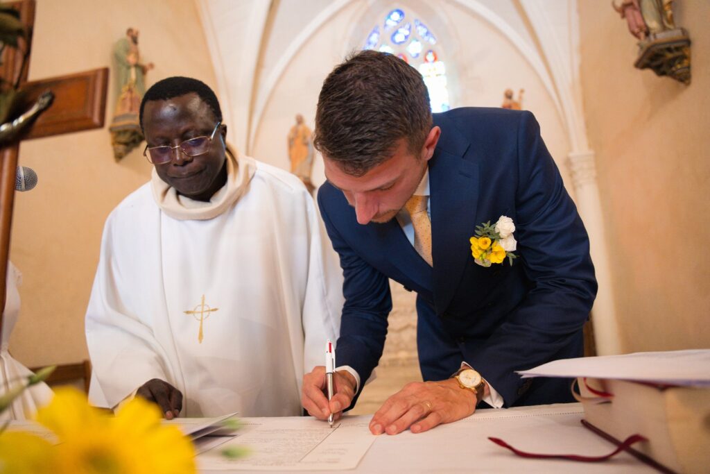 Un prêtre en aube blanche regarde le marié signer le registre sur l'autel, près d'un bouquet jaune.