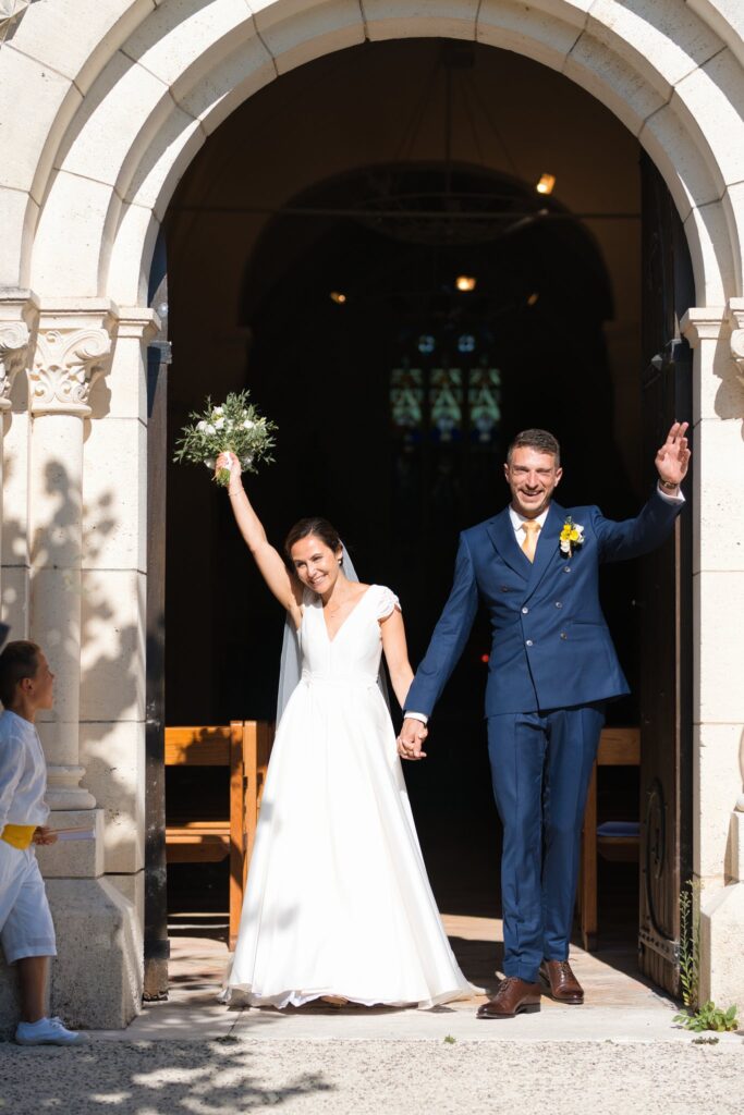 À la porte de l'église, les mariés sortent main dans la main, la mariée levant son bouquet sous une pluie de pétales.