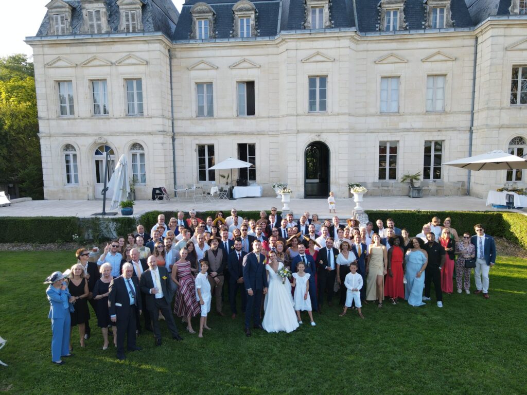Vue de drone : les mariés au milieu des invités rassemblés et saluant sur la pelouse devant le château.